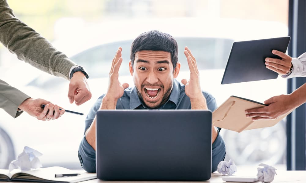 A frustrated man sits at a desk with his hands on his head staring with dismay at a computer screen as he is surrounded with people who demand his attention.