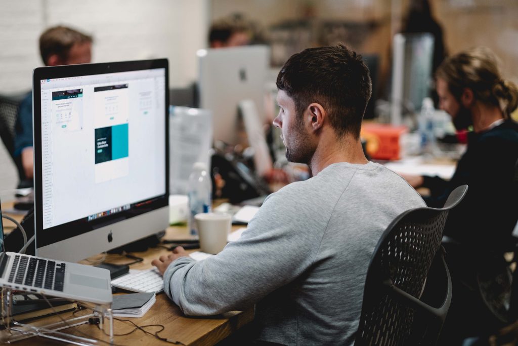 A man sits at a desk in an office, working on a large computer monitor displaying a design. Other workers are busy at their computers in the background. The workspace is filled with modern desks and office chairs.