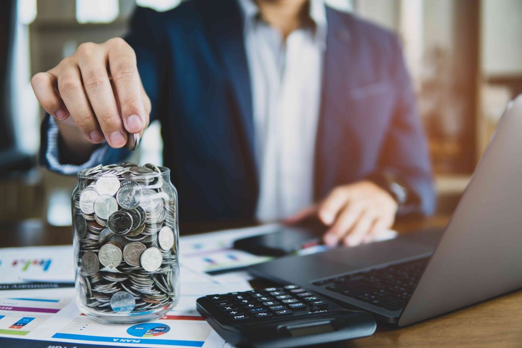 A person in a suit places a coin into a clear jar filled with coins, on a desk with paperwork, a laptop, a smartphone, and a calculator. The scene suggests financial planning or saving money.