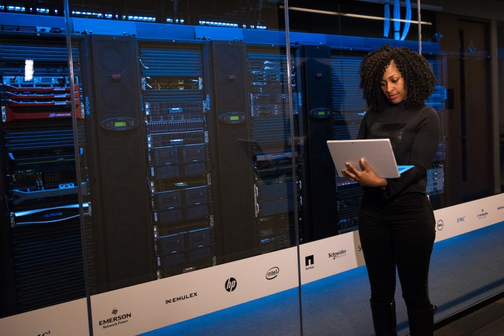 A woman stands in a server room, holding and looking at a laptop. Behind her are several tall server racks filled with equipment from various brands, visible through a glass partition. The room is dimly lit with blue-tinted lighting highlighting the servers.