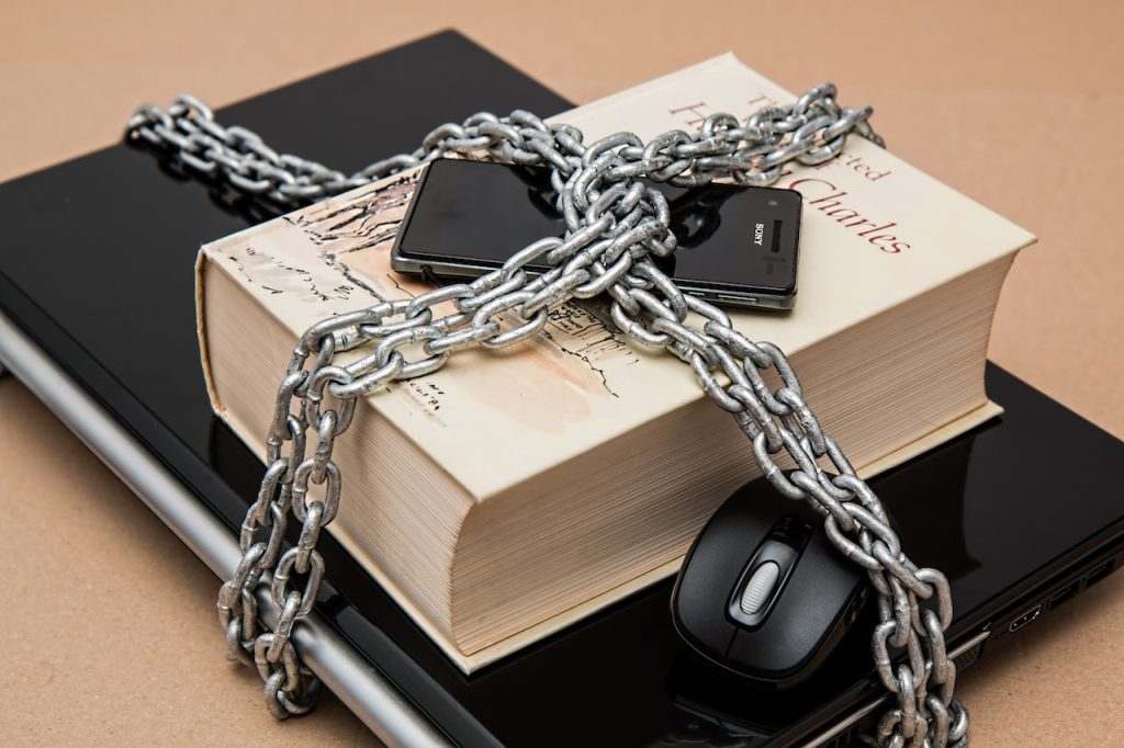 A book, two smartphones, and a computer mouse are placed on top of a laptop. All the items are tightly wrapped and secured with a thick metal chain, symbolizing restriction or confinement. The background is a plain, neutral surface.