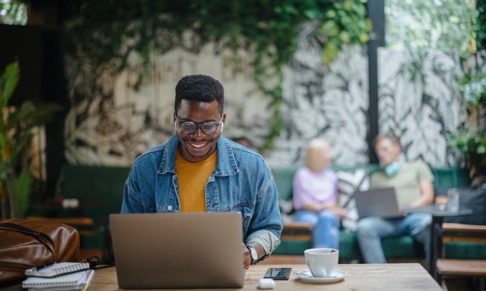 A person with glasses and a denim jacket is smiling while using a laptop in a cozy, plant-filled cafe. A notebook, pen, mouse, and coffee cup are on the table as they read about the best Microsoft Intune features. In the background, two people are sitting on a sofa, also working on laptops.