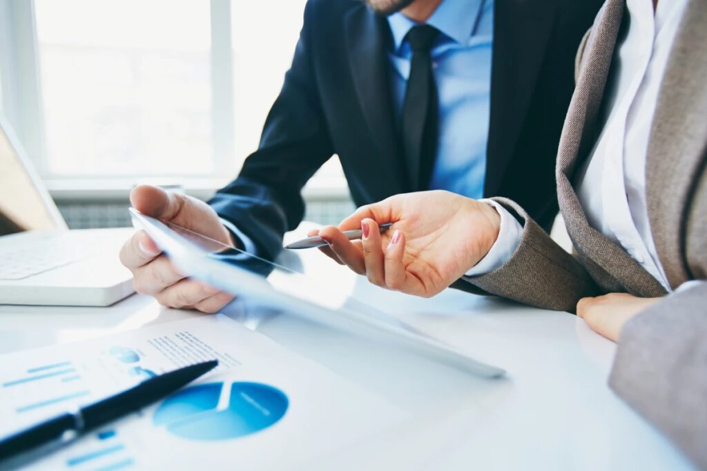 Two business professionals, partially visible, discussing a document on a tablet. A pen and financial graphs rest on the table in front of them. The focus is on their hands and the tablet, indicating a collaborative review or decision-making process at a Bellevue IT Company.