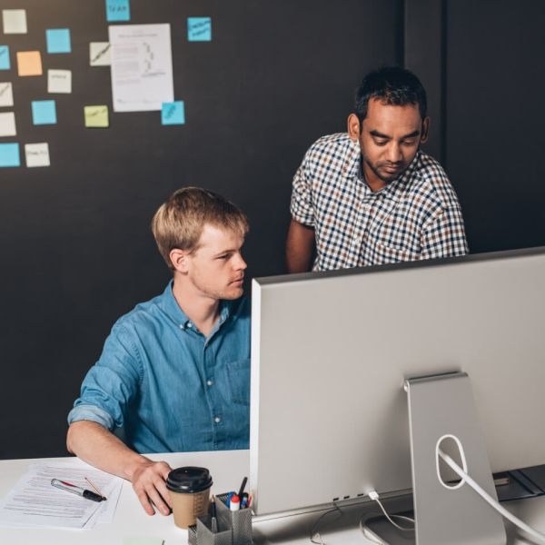 Two men collaborate in an office setting at their IT company in Bellevue. One, seated and focused on a computer screen, has a coffee cup nearby. The other leans over, pointing at the screen, seemingly giving advice. A dark wall behind them is dotted with colorful sticky notes and papers.