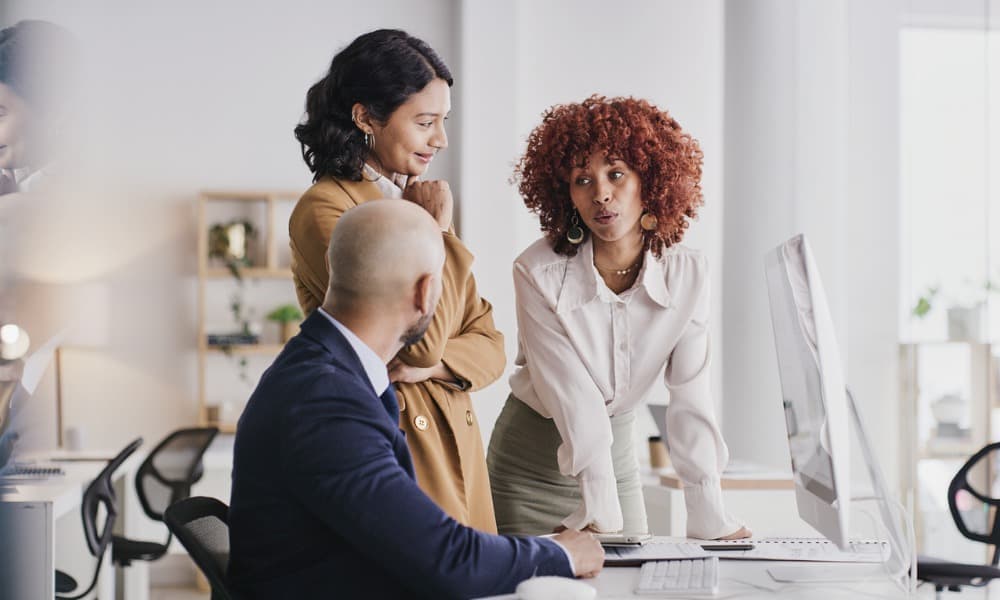 Three colleagues are engaged in a discussion at an office workstation. A woman with a curly hairstyle and a light blouse leans over the desk, sharing best intune practices with a man in a suit while another woman in a brown jacket and long hair stands nearby, listening attentively.