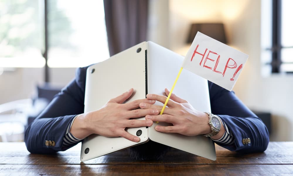 A frustrated businessman buries his head in a laptop while holding a sign that says help.