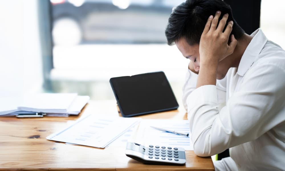 Stressed asian businessman holding his head confused by bills on desk.