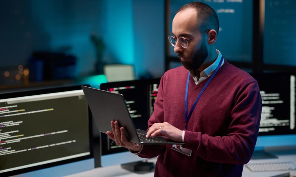 A man with glasses and a beard stands in an office, holding a laptop, fully focused on getting the most out of his work. Behind him, computer monitors display code against a blue-lit background, reflecting the tech-driven environment of his MSP partnership.