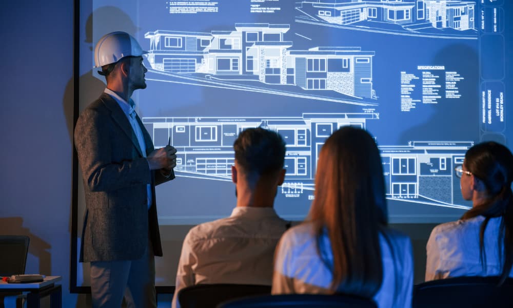 An engineer from a leading engineering firm presents architectural blueprints projected on a screen, outlining an infrastructure upgrade to an audience of three people seated in a dimly lit room.