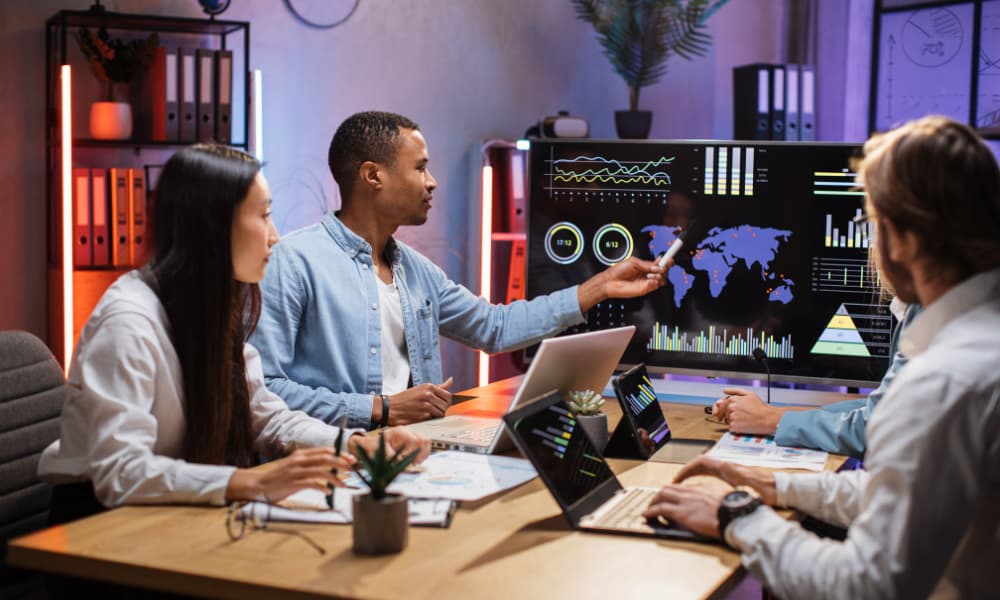 In a competitive modern office, a diverse team discusses data on a large screen showcasing I.T. trends with graphs and a world map. Laptops and documents on the table highlight their collaborative approach to navigating business challenges.