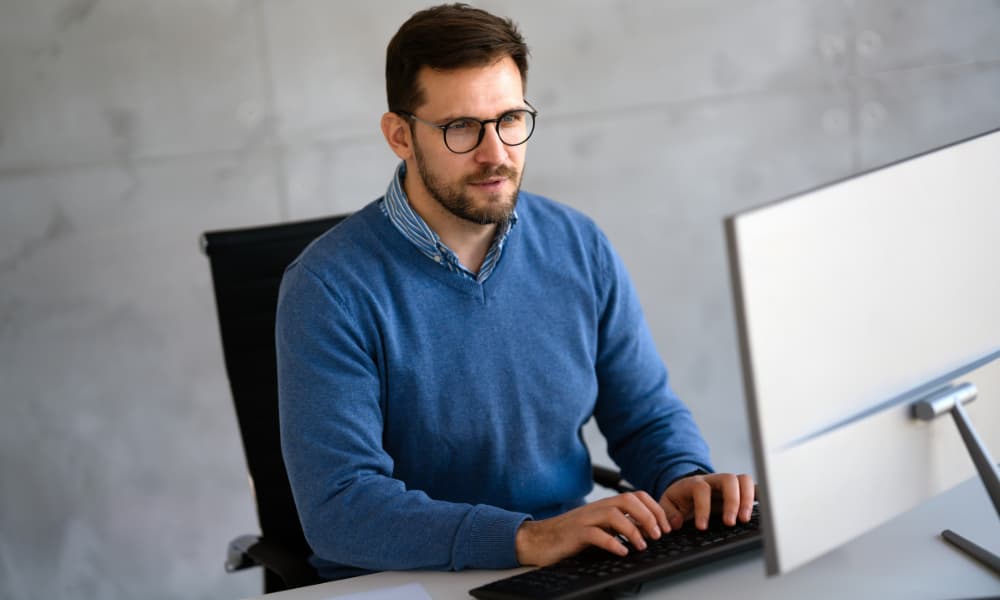 A man with a beard and glasses is at his desk, typing on a keyboard while focused on the computer monitor displaying Windows 10. He's sporting a blue sweater over a collared shirt, set against a plain, light gray wall.